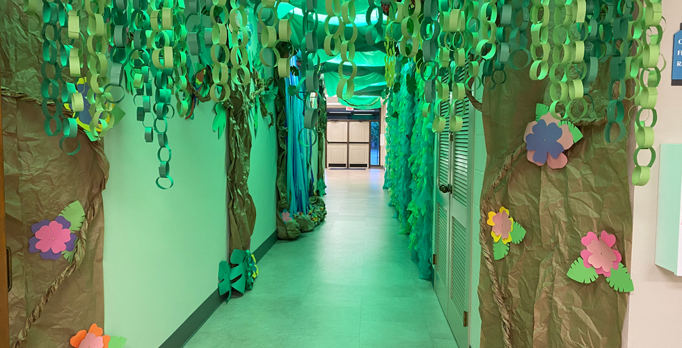 A long hallway with green paper chains and paper made to look like tree bark hanging in front. Down the hallway, some green tablecloth vines and a streamer waterfall can be seen. The hallway lights are green.