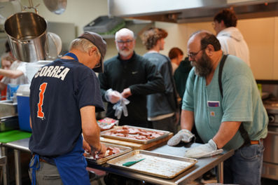 Three adult men stand around a table in the kitchen putting cut up sausage link pieces on pans