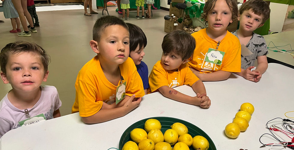 Six children stand at a table that has many wires and lemons on it