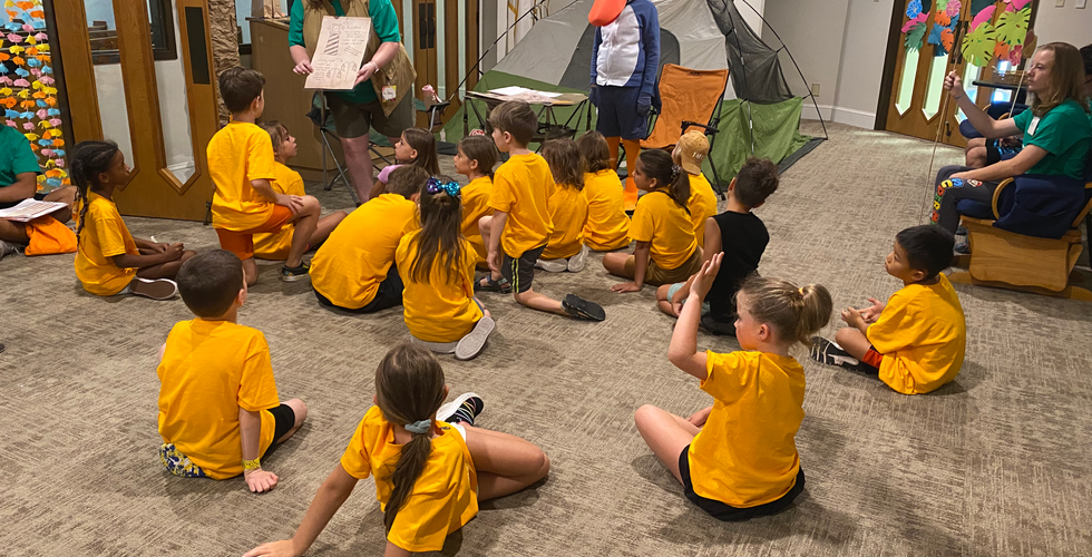 A group of children in yellow looking at an adult wearing adventurer gear holding up a piece of paper with a lighthouse on it
