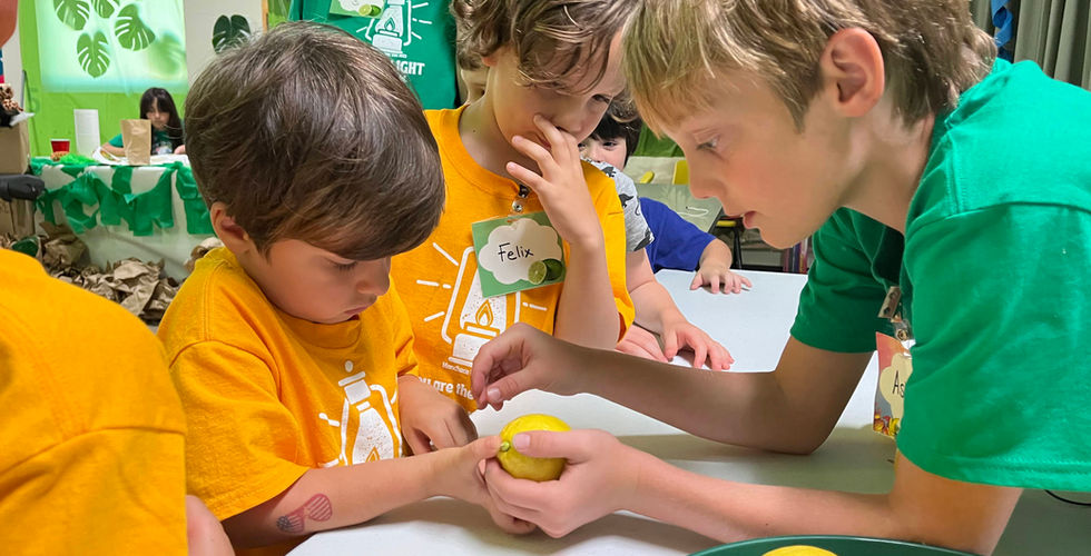 A youth boy in a green shirt helps a little boy in a yellow shirt put a penny into a slit in the lemon over a table. A few other children are surrounding them.