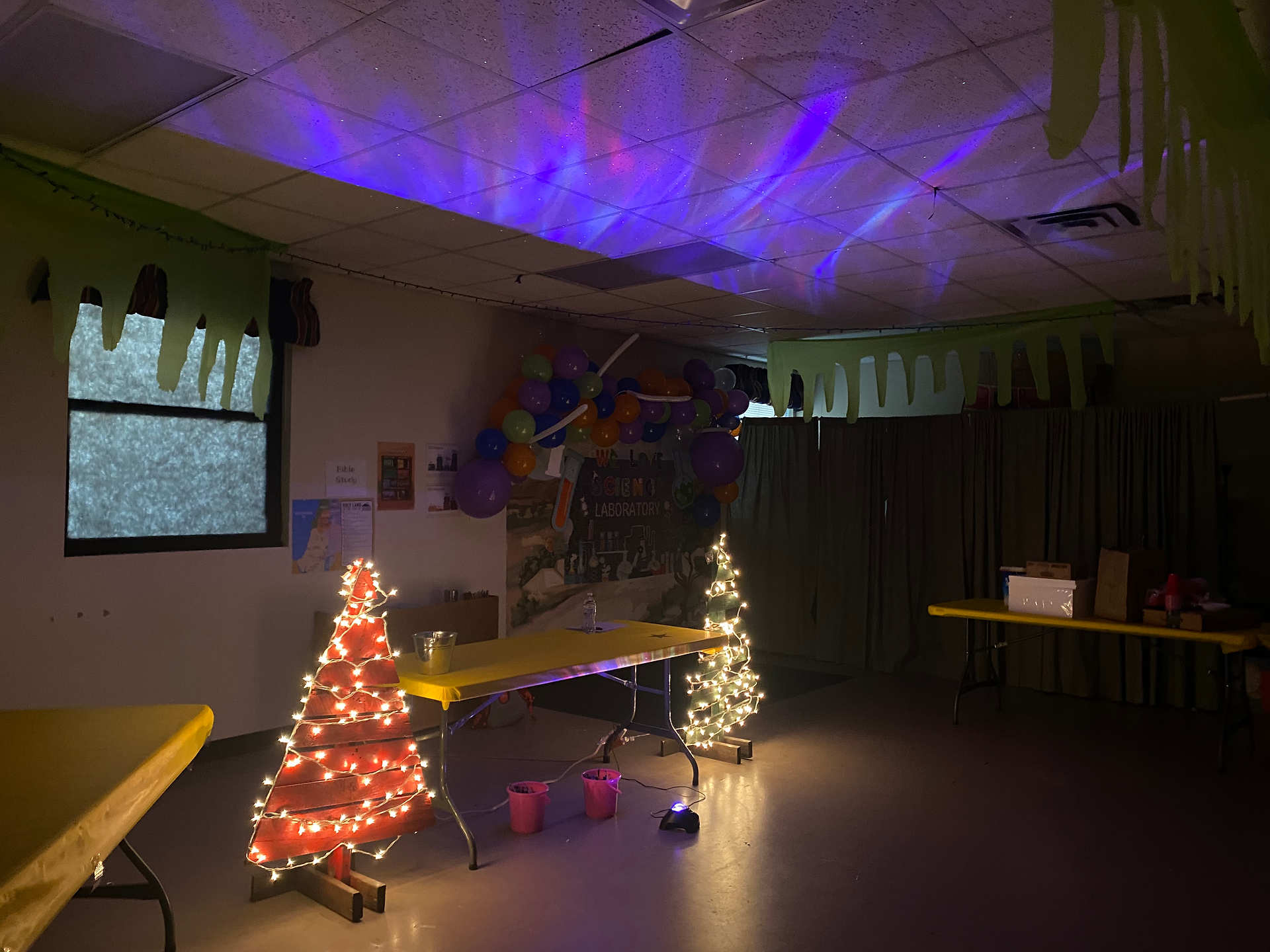 A classroom decorated with a few light-up Christmas trees, an aurora borealis lights projected on the ceiling, and more tablecloth to look like slime.