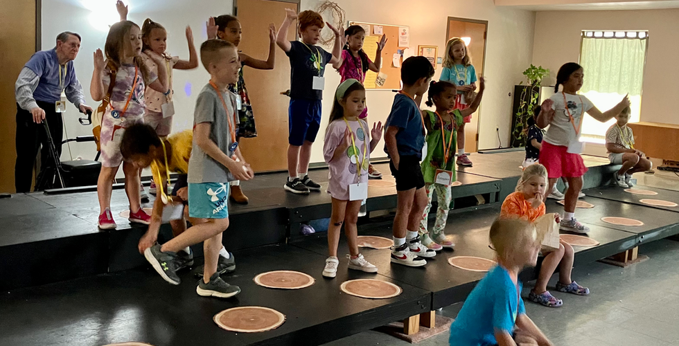 A group of children on black risers are in the middle of a dance with their arms up.