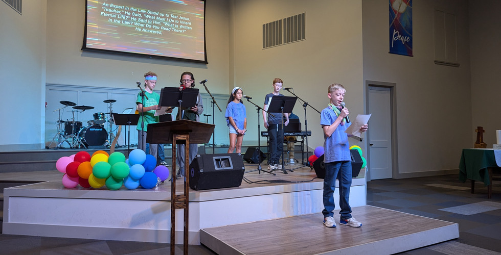 A teenage boy is standing on the lower stage reading off a piece of paper into a microphone. Behind him is a taller stage where four other teenagers are standing with a man playing guitar and a man playing piano.