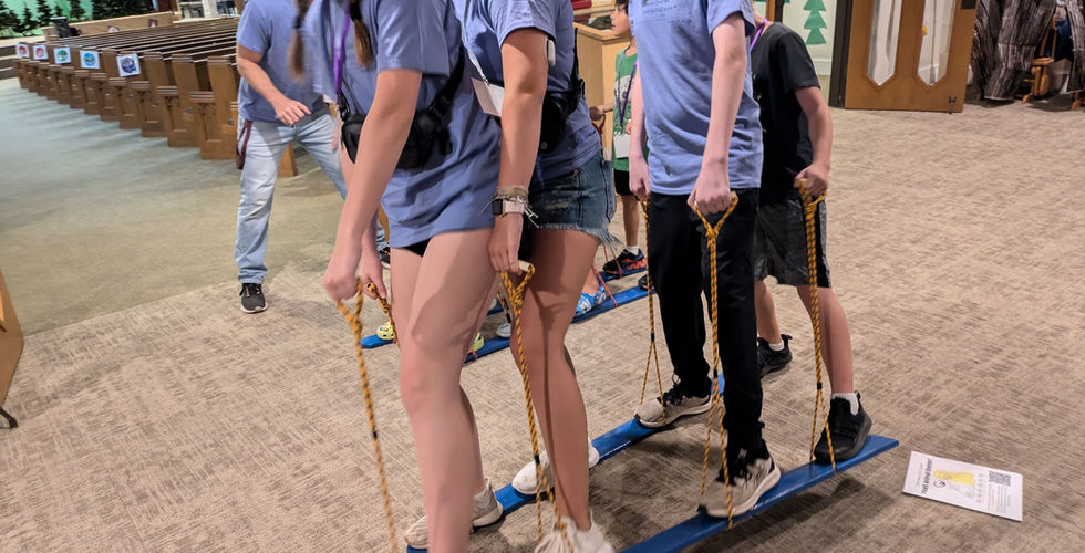 Three teenagers in blue shirts and a child, whose face is hidden, doing the plank walk activity. Their left feet are in the air with the board successfully held up.