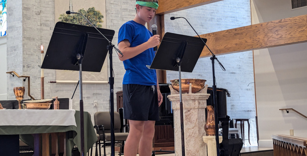 A teen boy is reading off of a music stand and speaking into a microphone.