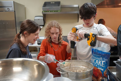 Three teenagers stand around a pot filled with pancake batter. The teen in the middle is holding a spoon over the bowl, and the one on the right is holding a power drill with a long stick to stir it.