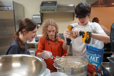 Three teenagers stand around a pot filled with pancake batter. The teen in the middle is holding a spoon over the bowl, and the one on the right is holding a power drill with a long stick to stir it.