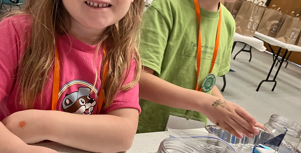 A young girl and a young boy are standing in front of metal trays with a bottle of blue liquid and a little boat inside in front of them. The young girl is smiling at the camera and the young boy is in the middle of talking.
