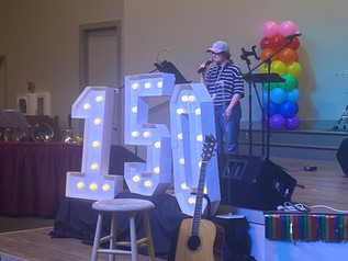 A woman is singing into a microphone on a stage behind big 150 numbers