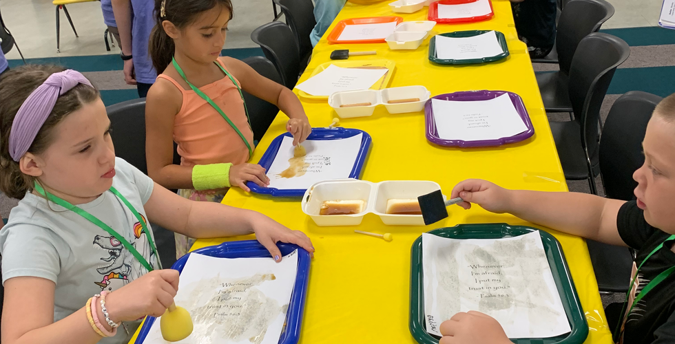 Children sitting around a rectangular table covered in yellow tablecloth. There is a tray in front of each kid with a piece of paper on it, and they are dabbing the paper with coffee.