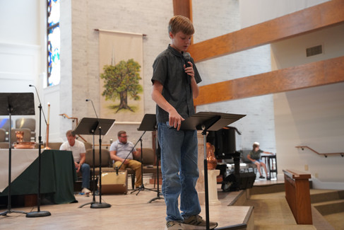 A teen in a grey button up shirt and jeans talks into a microphone at the front of the sanctuary