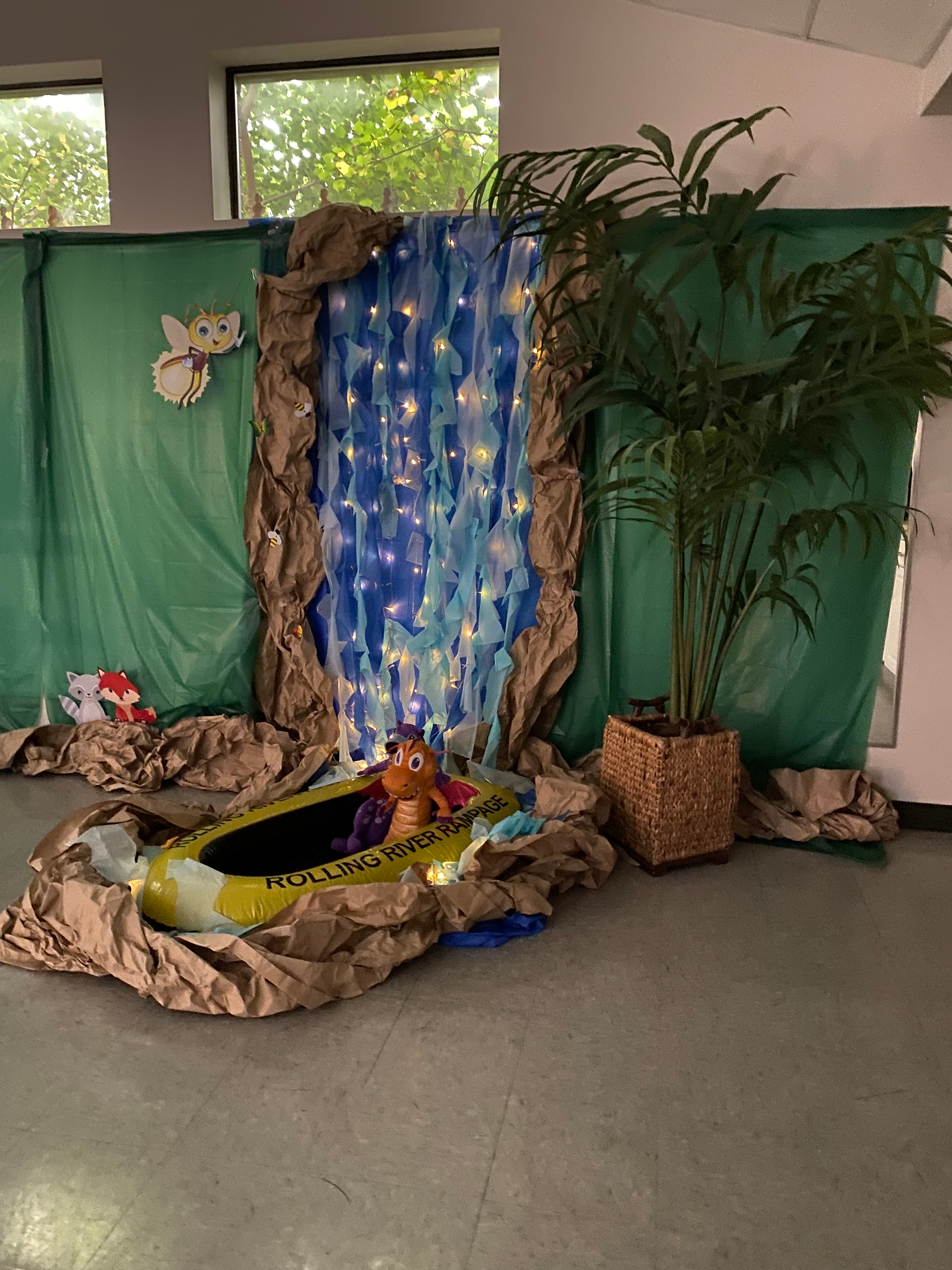 A waterfall made out of tablecloth attached the wall. There is a little pond at the end of the waterfall that has a raft with a stuffed dragon and an otter inside. There is green paper behind the waterfall with some animals taped to it, and a big fake tree to the right.