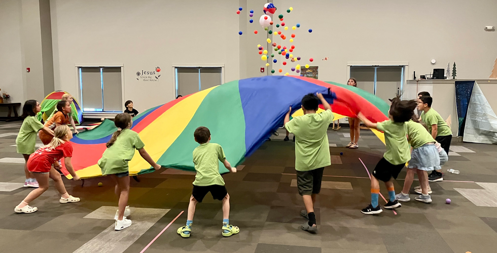 A group of children and teens stand around a colorful parachute in varying degrees of lifting. There are a ton of little colorful balls flying up above the parachute.