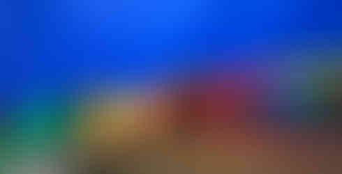 A line of children are sitting on the edge of the colorful parachute inside of it. In the middle of the photo is an adult woman taking a photo with one of the children.