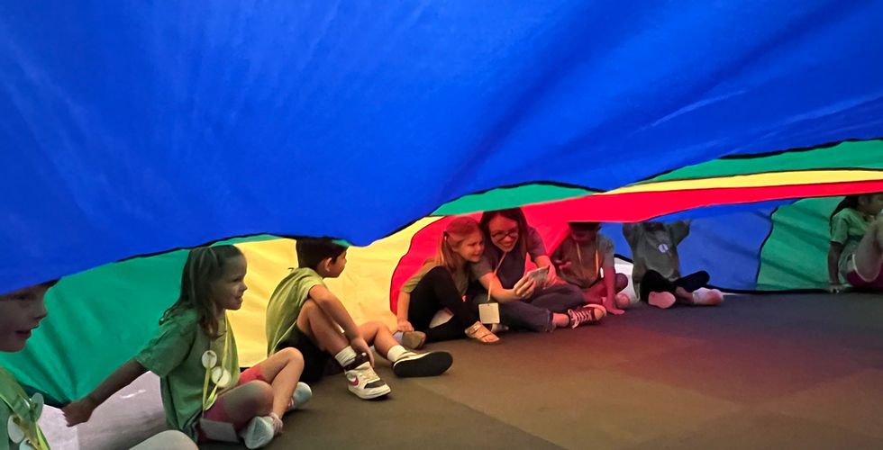 A line of children are sitting on the edge of the colorful parachute inside of it. In the middle of the photo is an adult woman taking a photo with one of the children.