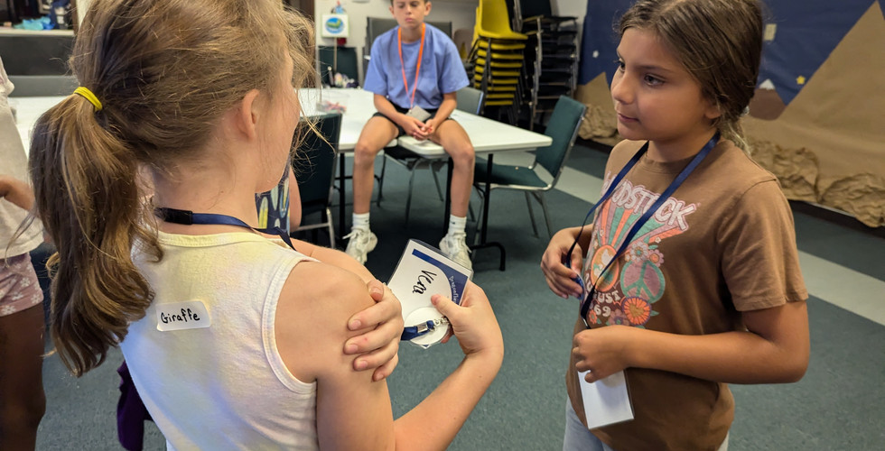 Two young girls stand facing each other. The girl facing away from the camera has a label on her back with the word "giraffe" on it.