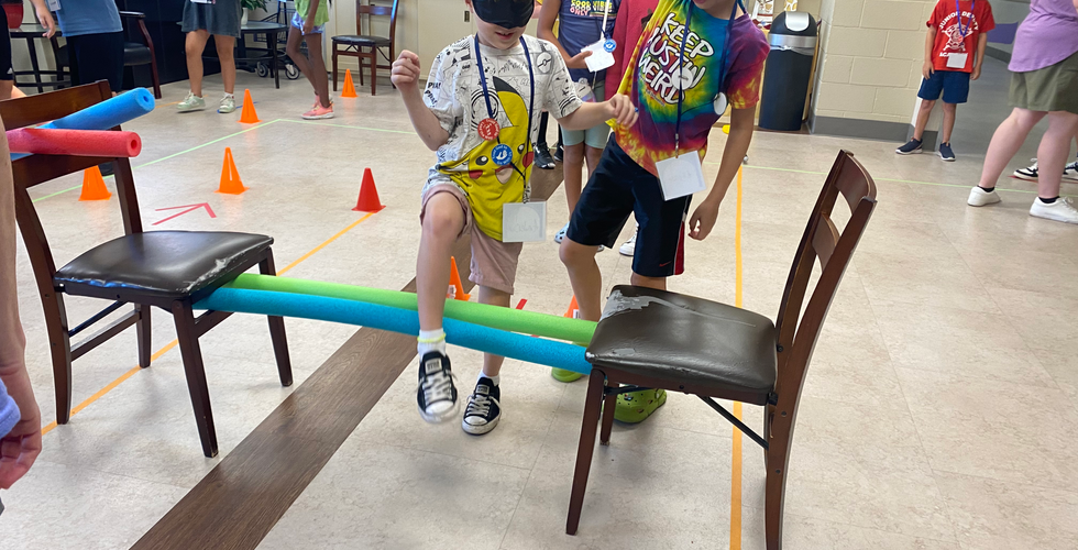 A child in a white and yellow shirt is blindfolded and stepping over two pool noodles propped up on chairs. A young boy to the right of him is holding his arm up and trying to guide him over the pool noodles.