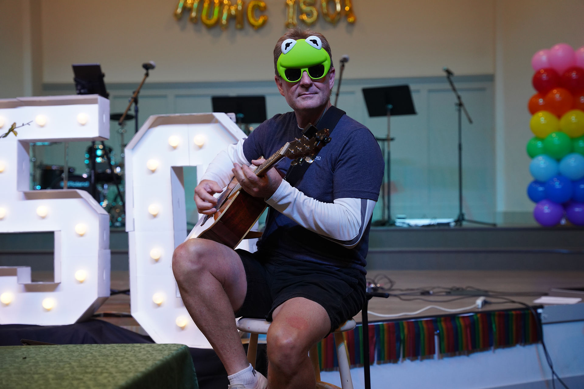 An adult man sits on a stool playing piano. He is wearing sunglasses with Kermit the Frog on them.
