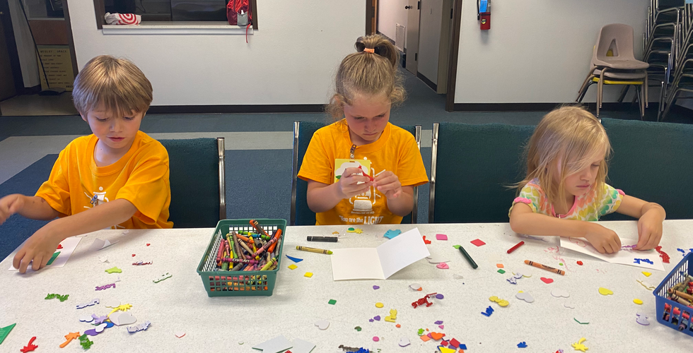 Three children sit at a table coloring and putting stickers on cards