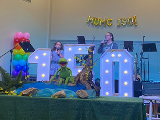 A young girl and a teenage boy singing into microphones on a stage behind a display of Kermit the Frog and big 150 numbers