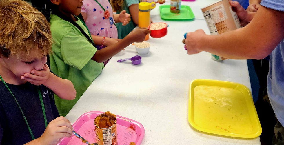 Children are lined on up the left side of the table with adults on the right. The child closest to the camera is spooning pumpkin filling into a measuring cup and covering his mouth in disgust.