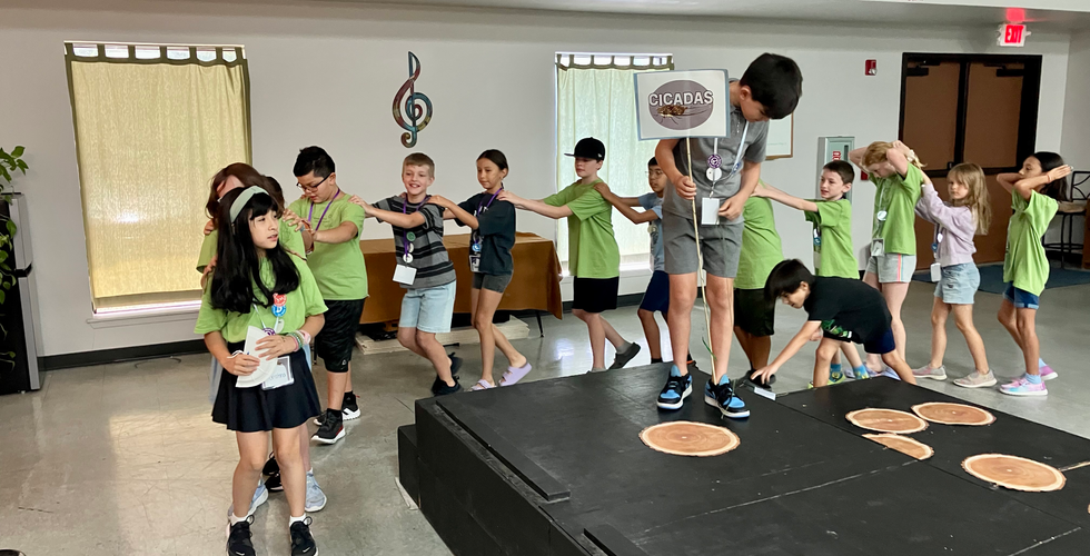 A line of children walking around black risers in a conga line. There is a child on the risers holding a flag that reads "Cicadas."