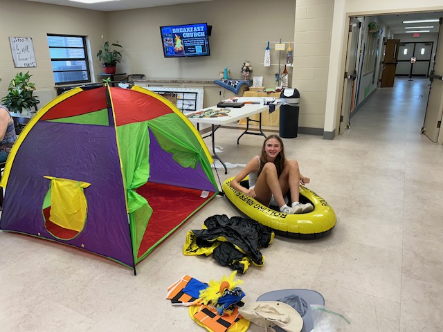 A teen girl sits in a small yellow blow-up raft in an open room with a colorful tent behind her and camping supplies around her.