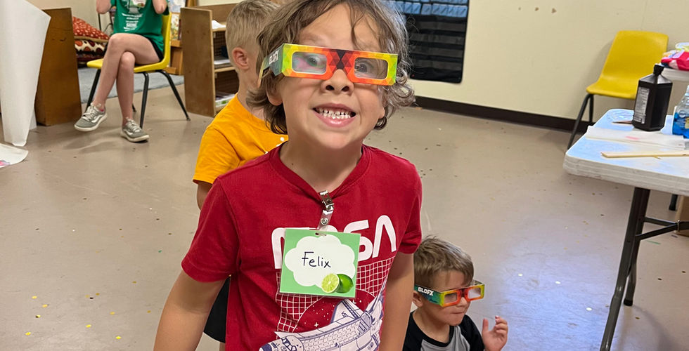 One young boy in a red shirt smiles at the camera wearing multicolor paper glasses that make light appear crystalized.