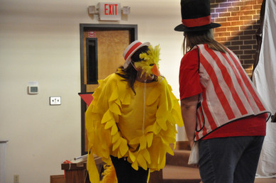 A woman dressed up as a yellow bird and a second woman facing away from the camera with a red and white striped vest and a top hat. Both are bent over as if bowing to each other.