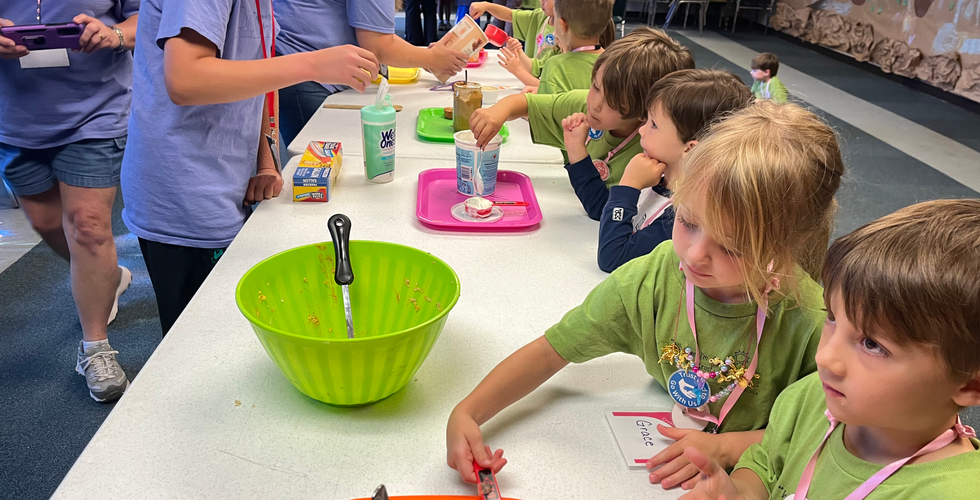 Children in mostly green shirts stand on one side of a table with adults in blue shirts stand on the left. There are trays and bowls with ingredients along the table, as some of the children are pouring things into their containers.