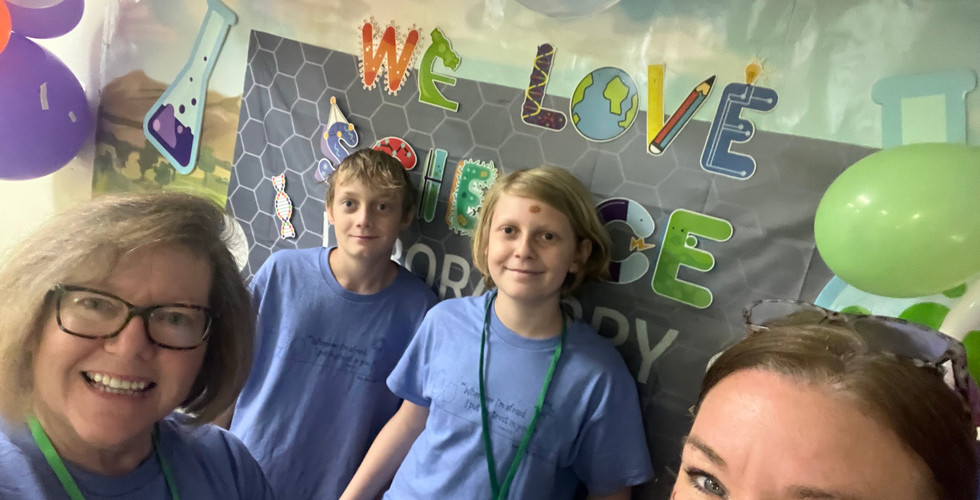 Two adult women and two teen boys stand in front of a photo display that says "We love science" and some balloons.