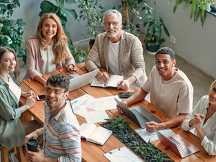 Grupo diverso sorrindo em reunião em sala com muitas plantas, laptops, cadernos e gráficos sobre mesa de madeira.