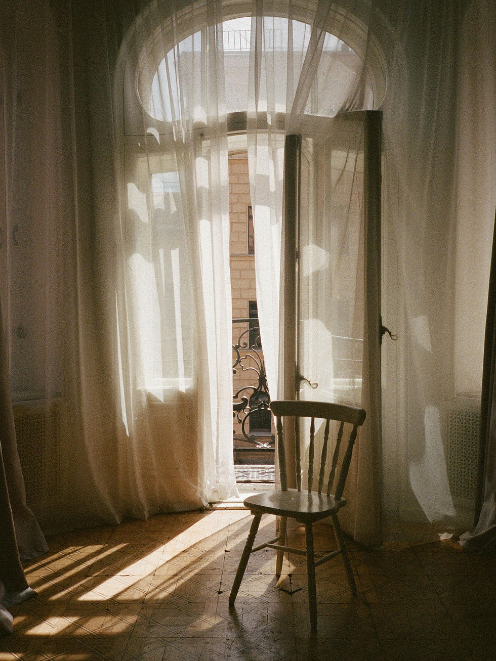 Grande Janela de sacada com feixe de luz passando pela cortina e refletindo no chão. No centro à frente, uma cadeira vazia
