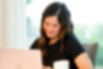 Smiling woman with long dark hair works at a pink laptop beside a white mug in a bright home office.