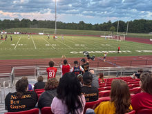 Fans celebrate a Hub City FC goal by standing, cheering, and waving scarves.