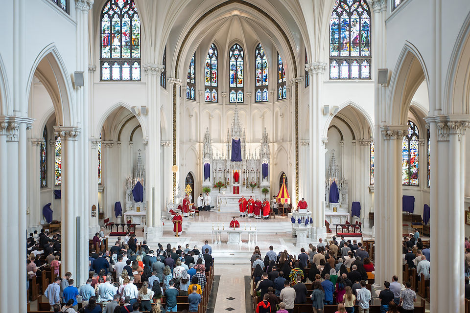 Large congregation in a cathedral with stained glass, arches, and clergy in red robes at the altar. The scene is solemn and reverent.