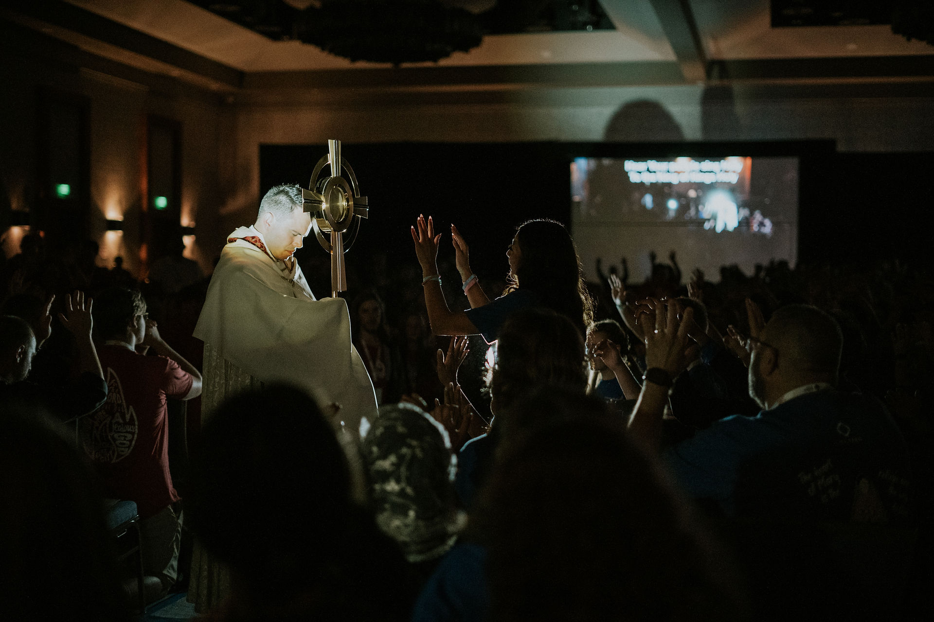 Priest holding a monstrance, surrounded by people with raised hands in a dimly lit room. Projected text visible on the background screen.