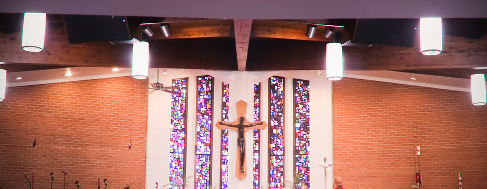Church sanctuary with congregation seated in pews, altar beneath crucifix and stained-glass windows, a man in red speaking at lectern