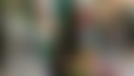 Priest in green vestments speaks into a microphone outdoors, with colorful pillar and greenery in the background. Calm and reflective mood.