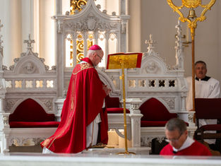 Obispo católico de rojo orando ante un atril en una iglesia ornamentada, con ayudantes al fondo y ambiente solemne.