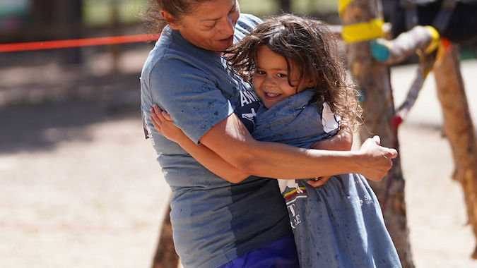 Woman joyfully spins a child in her arms outdoors. Both wear blue shirts. Background has trees, colorful ropes on poles, and a sandy ground.