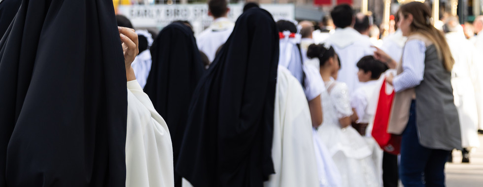 Nuns and children in white and black robes walk in a city street crowd near a religious procession, with tall buildings behind.