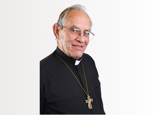 Elderly man in clerical attire with a cross necklace, smiling against a plain white background, conveying a warm, calm mood.