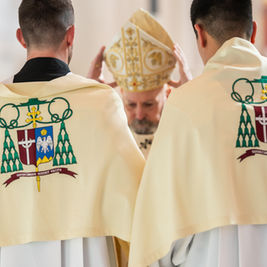 Dos personas con capas bordadas, de espaldas, frente a un hombre con mitra dorada levantando las manos. Interior de iglesia. Ambiente solemne.