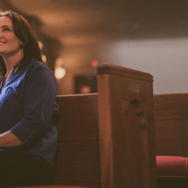 Woman in blue shirt sitting in a church pew, smiling softly. Warm lighting with wooden pews and blurred background create a serene mood.