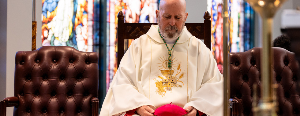 Seated clergy in ornate white robes holds a red cap, eyes closed in prayer. Stained glass windows create a colorful backdrop.