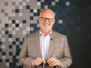 Smiling man in glasses and plaid jacket stands against a patterned pixelated background, conveying a cheerful mood.