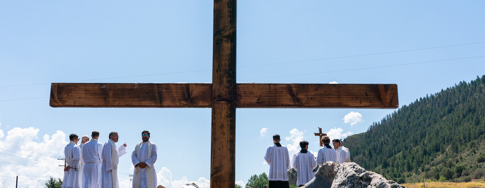 White-robed priests gather outdoors around a large wooden cross, with mountains and blue sky behind them, in a solemn ceremony.