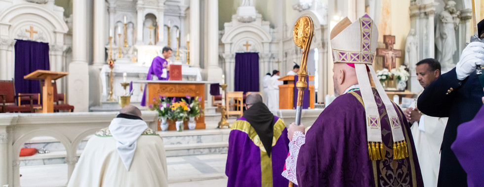 Bishops in purple robes stand in a church, one holding a staff, with altar and floral arrangements in the background. Stained glass visible.